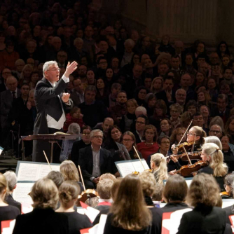 Andrew Carwood, Director of Music, conducts an orchestra with a packed audience in the background.