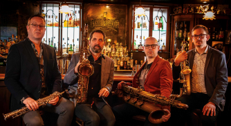 A man and three women holding string instruments sitting on a bench in front of a stone window