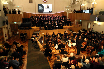 An audience applauding the end of a concert, featuring a conductor, orchestra and large choir on stage.